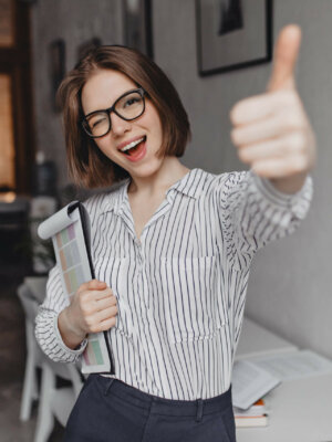 Young woman in office style clothes and glasses holds tablet with documents, winks and shows thumb up